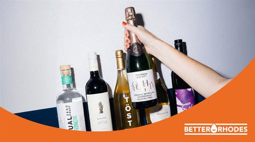 A hand holding a non-alcoholic sparkling wine bottle surrounded by alcohol-free spirits and wines