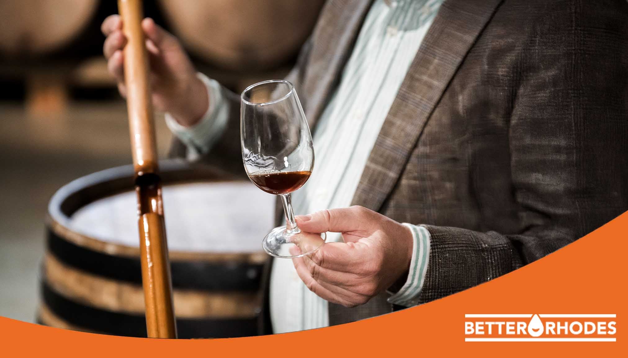 Person holding a wine glass and tasting a beverage inside a barrel room